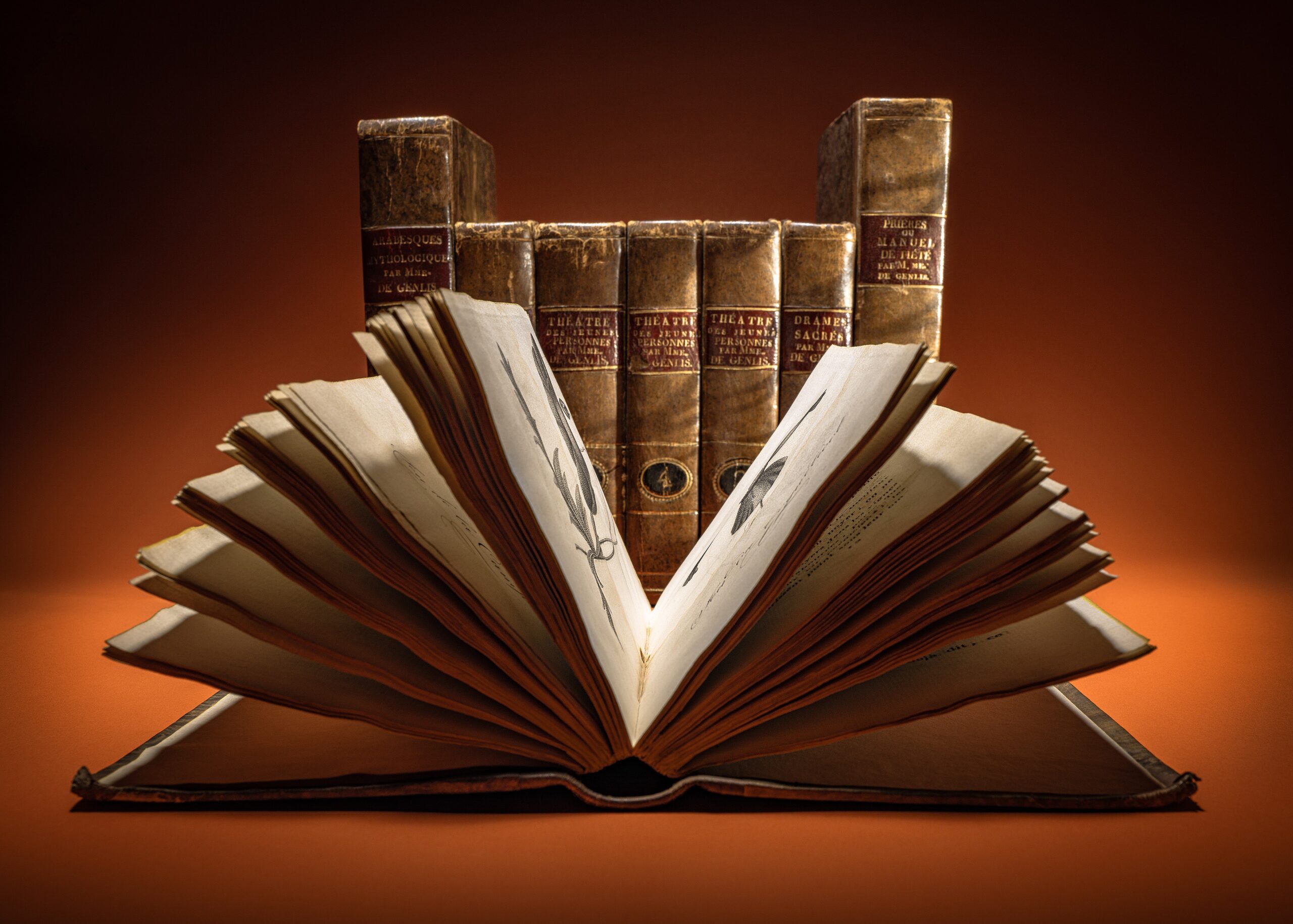 Een oud boek opengeslagen met op de achtergrond de rug van 7 oude boeken.