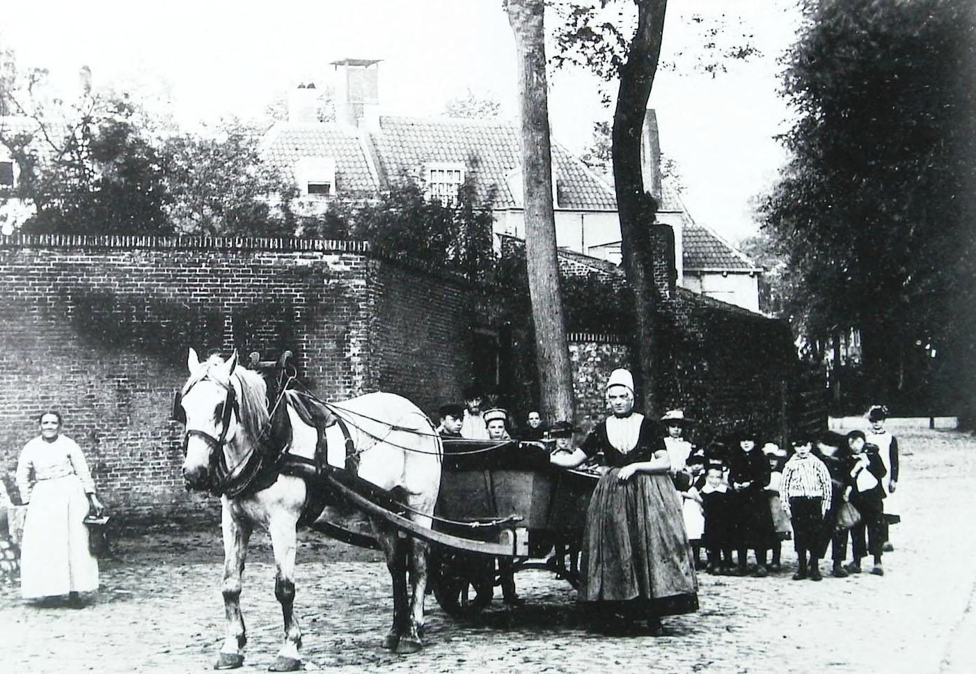 Een zwart-wit foto van een vrouw in klederdracht, een melkboerin, die haar hand op haar wagen legt en in de camera kijkt. De wagen wordt getrokken door een paard. Achter de wagen staat een grote groep jonge kinderen. Links op de foto staat een glimlachende vrouw met een mandje in haar hand. Links op de achtergrond is een muur te zien met daarachter bomen, daken en schoorstenen. Rechts zijn ook bomen te zien.
