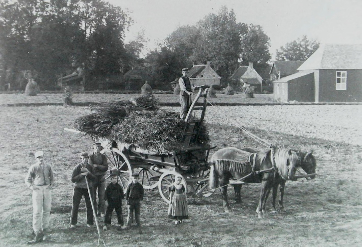 Een zwart-wit foto van vier mannen en drie kinderen op het land. Eén man staat boven op een met landbouwproducten volgeladen kar die wordt getrokken door twee paarden. De drie andere mannen staan links op de voorgrond, een van hen leunt op een stuk gereedschap. Naast hen staan twee jongetjes en een meisje in dracht. De foto is duidelijk geposeerd: alle personen kijken naar de camera. Op de achtergrond zijn wat bomen, kleine huisjes en, wat lijkt op een boerderij te zien. Ook staan er strobalen in het veld.