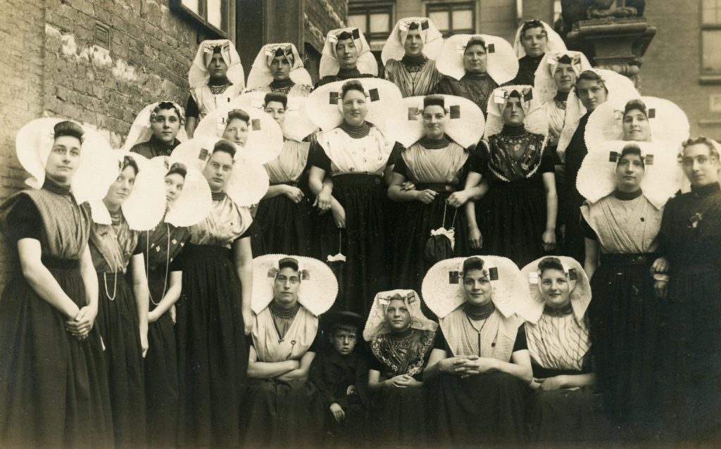 Een groep dames in Zuid-Bevelandse klederdracht met in het midden Prinses Juliana. De meeste vrouwen dragen een witte schelpvormige muts van kant: zij zijn protestants. De katholieke vrouwen dragen een muts met een hoekig, trapeziumachtig model. Alle mutsen zijn voorzien van vierkante gouden 'stukken'.