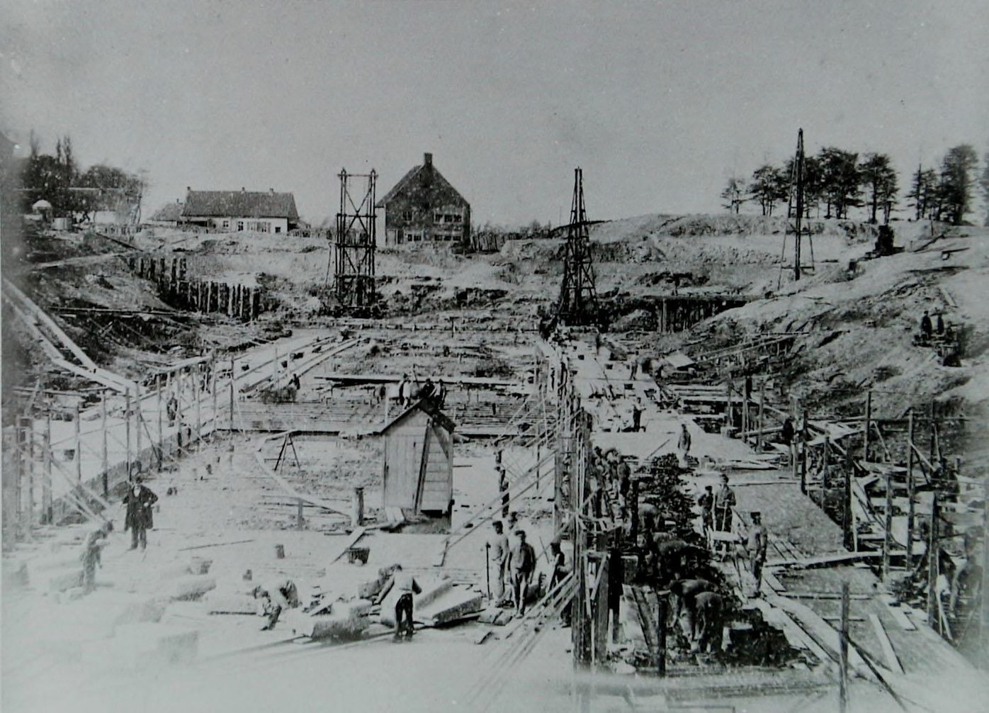 Hele oude zwart-wit foto met op de achtergrond enkele huizen en bomen, in het midden enkele werktuigen en op de voorgrond een grote groep mannen die aan het werk is in het droogstaande kanaal.