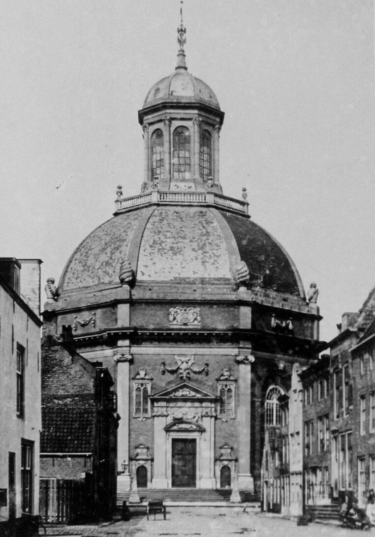 Zwart-wit foto van een achtkantige, barokke koepelkerk. De foto is genomen op zo'n 50 meter afstand van de kerk waardoor je aan beide kanten huizen ziet. De voorkant van de kerk heeft een trap die naar de ingang leidt, voor de ingangstrappen op het twee hardstenen obelisken. Op het koepel dakstaat een rond torentje met hoge ramen, daar bovenop staat nog een koepeldakje. Voor de volledige omschrijving: https://monumentenregister.cultureelerfgoed.nl/monumenten/29376