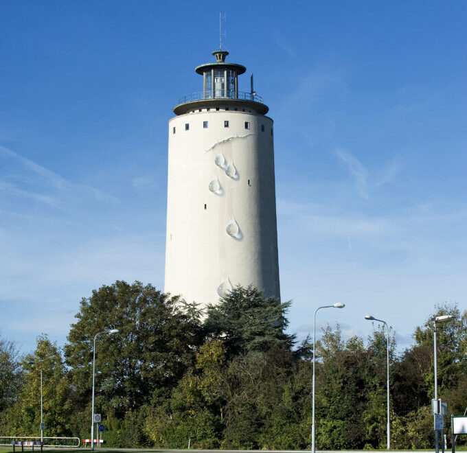 Een witte watertoren met daarop kunstwerk "De Bron", bestaande uit een geschilderde scheur en waterdruppels, waardoor het lijkt alsof er water uit de toren stroomt. Op de voorgrond bomen.