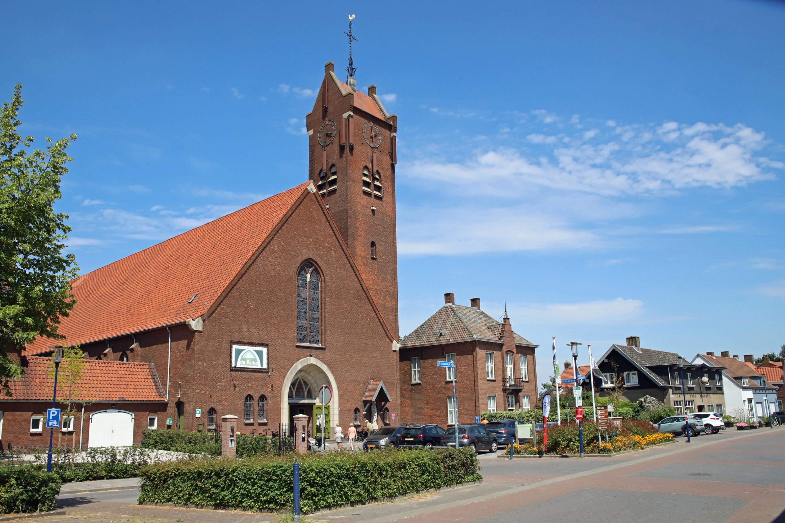 Rood bakstenen kerkgebouw met hoge klokkentoren en uurwerken aan een dorpsstraat, onder een helderblauwe lucht. Voor de kerk staan enkele mensen en geparkeerde auto’s.